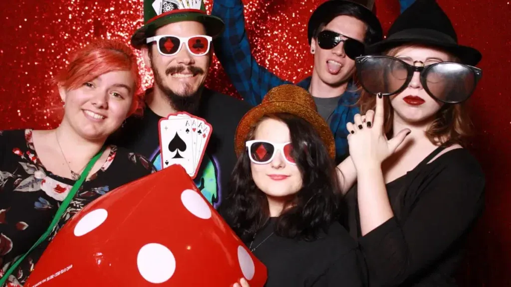 students wearing casino card glasses, holding dice, and posing in front of a sparkly red background